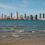San Diego skyline with modern skyscrapers seen from across the bay on a clear day, gentle waves in the foreground, and blue sky above—a perfect backdrop for a round on a golf simulator or an afternoon stroll.