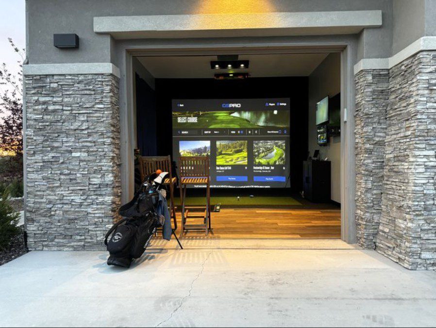 A home garage converted into a golf simulator, with a golf bag and clubs in the foreground and a large screen displaying golf graphics on the back wall. Two chairs face the screen, and the garage has stone accents.