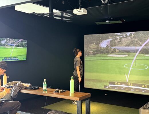 Two people are in a golf simulator room. One stands near a large screen displaying virtual golf stats and a course, while the other sits on a bench using a phone. Golf clubs and water bottles are on the table.
