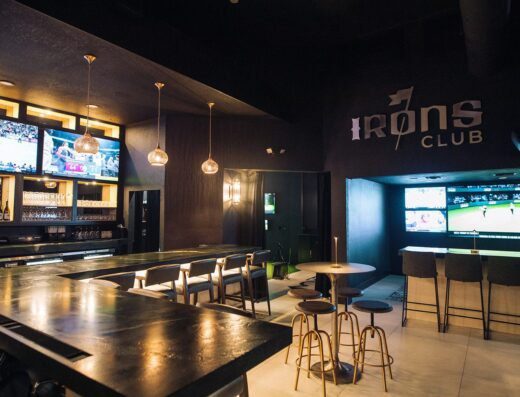 Modern sports bar interior with a long bar counter, barstools, and several flat-screen TVs displaying sports. The wall features the IRONS CLUB logo. The space is dimly lit with hanging pendant lights.