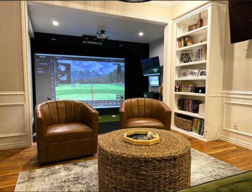A cozy room with two brown leather chairs, a round wicker coffee table, and a golf simulator setup with a large screen showing a golf course. Built-in shelves hold books and decor on the right.