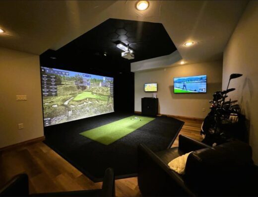 A modern indoor golf simulator room with a large screen displaying a golf course, an artificial turf mat, a golf bag, a wall-mounted TV showing golf, and comfortable seating under warm recessed lighting.