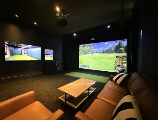 A modern indoor golf simulator room with a large screen showing a golf course, two flat-screen TVs on the wall, golf clubs, and a seating area with leather sofas and a wooden coffee table.