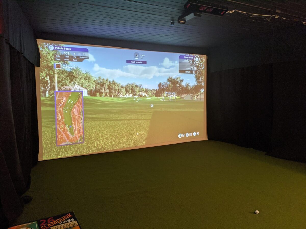 A golf simulator room with a large screen displaying a golf game, green turf flooring, a golf ball on the floor, and part of a sign visible in the lower left corner.
