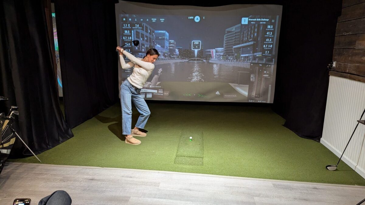 A person in jeans and a white sweater swings a golf club indoors on artificial turf, facing a virtual golf simulator screen displaying a night cityscape with data.