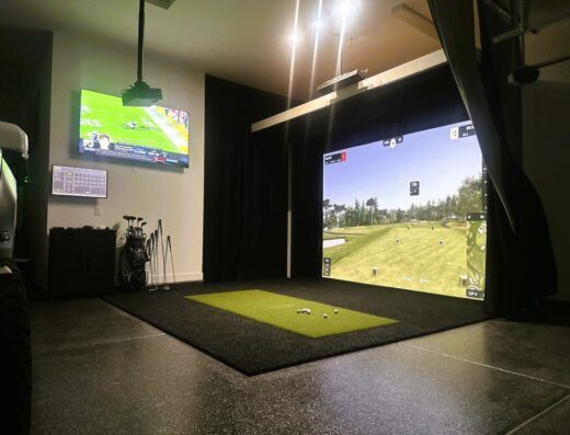 A home golf simulator setup with a large projector screen displaying a golf course, artificial turf mat, golf clubs nearby, and a TV on the wall showing a football game. Bright overhead lights illuminate the space.
