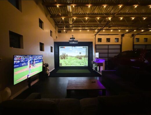 A modern garage features a golf simulator setup with a large screen showing a golf course, a mounted projector, a TV displaying a baseball game, a couch, and string lights hanging from the ceiling.