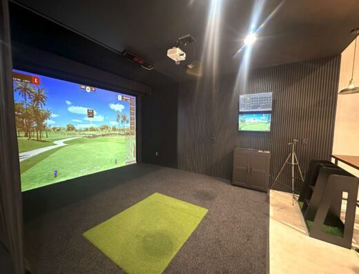 A modern indoor golf simulator room features a large screen displaying a golf course, a green mat on the floor, black walls, a projector on the ceiling, and a monitor mounted on the side wall.