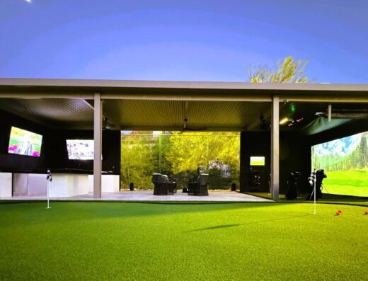 A modern outdoor golf simulator setup with large digital screens, golf clubs, and artificial turf, illuminated under a night sky, surrounded by greenery in the background.