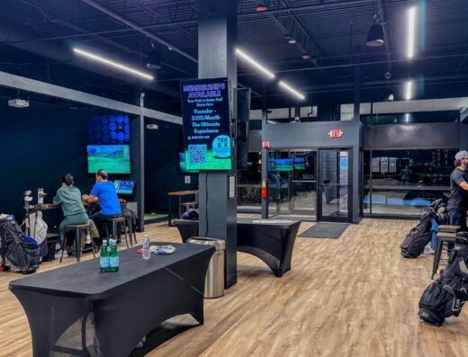 Indoor golf simulation facility with people playing at golf simulator stations, golf bags on the floor, tables with drinks, and a large digital sign displaying information. The space has wood flooring and modern lighting.