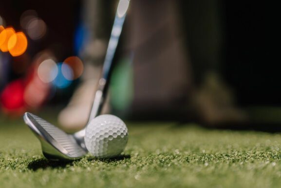 A close-up of a golf club and golf ball on green artificial turf, ready for a shot, with blurred lights and a golfer’s shoes visible in the background.